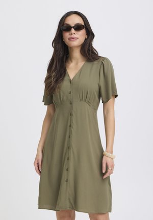 Kobieta ubrana w oliwkowo-zieloną sukienkę na guziki, czarne okulary przeciwsłoneczne, złote kolczyki i kremową bransoletkę, stojąca na tle jednolitego jasnego tła.