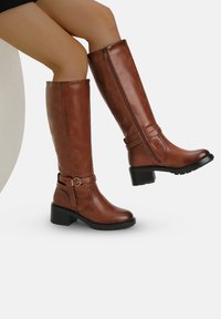 Botas marrones hasta la rodilla, hechas de cuero sintético, con un cierre lateral, una correa con una hebilla circular y un tacón grueso.