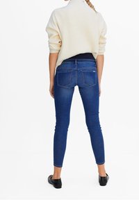 Jeans ajustados de mezclilla azul con una cintura alta, diseño de cinco bolsillos y un ligero estiramiento, combinados con un suéter de punto crema y zapatos negros.