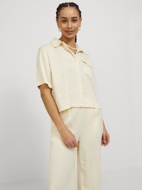 Camicia crema, a maniche corte, con bottoni, realizzata in tessuto liscio, caratterizzata da una tasca sul petto e un orlo leggermente accorciato, abbinata a pantaloni coordinati.