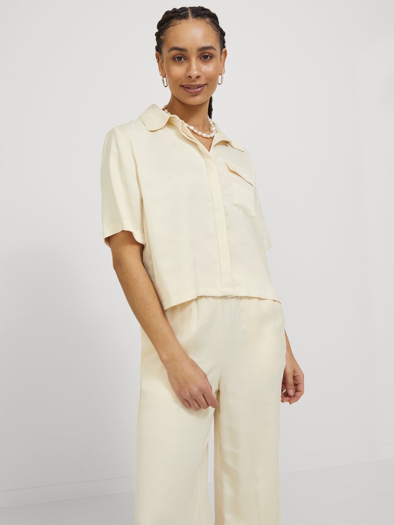 Camicia crema, a maniche corte, con bottoni, realizzata in tessuto liscio, caratterizzata da una tasca sul petto e un orlo leggermente accorciato, abbinata a pantaloni coordinati.