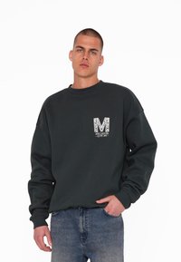 Tmavo zelená mikina s rebrovanými manžetami, okrúhlym výstrihom a bielym grafickým logom "M". Nosená s svetlomodrými džínami.
