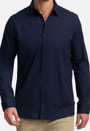 Man wearing a fitted navy blue long-sleeve button-up shirt with beige pants, standing against a plain white background.