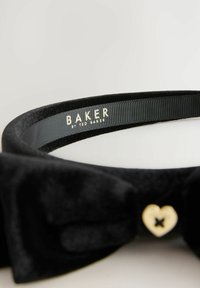 Fascia in velluto nero con un grande fiocco e un accento a forma di cuore dorato con una X nera. Branding visibile sulla parte interna della fascia.