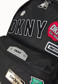 Black nylon backpack featuring bold embroidered "DKNY" lettering, various colorful patches, and metal label accents with brand details.