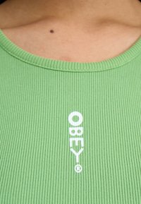 Grønn ribbestrikket topp med bred halslinning som har hvit "OBEY" tekst. Materialet ser mykt ut med en glatt tekstur og lett stretch.