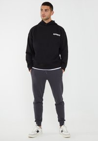 Sudadera con capucha negra con el logo "ASPORTO", combinada con joggers grises y zapatillas blancas. Textura suave, ajuste relajado, cordón ajustable.