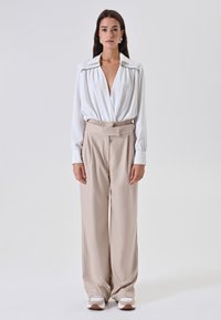 Blusa bianca a maniche lunghe con scollatura a V profonda e dettagli con bottoni, abbinata a pantaloni beige a vita alta e a gamba larga. Tessuto liscio, vestibilità rilassata.