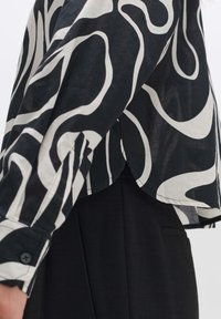 Camicia a motivi in bianco e nero con un design cropped, caratterizzata da linee fluide e ondulate. Maniche lunghe con polsini abbottonati, realizzata in tessuto leggero.