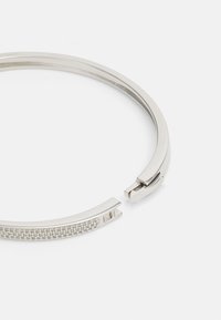 Bracelet ton argent avec fermoir à charnière et détail de surface rectangulaire texturée sur un côté, présenté partiellement ouvert sur fond blanc.