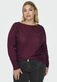 Bordo rebrasti pulover z barko izrezom in dolgimi rokavi, v kombinaciji s črnimi hlačami, ki imajo dva zlata gumbka na pasu.