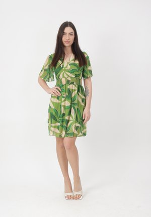 Vrouw met lang donker haar in een korte groene jurk met bladmotieven, poseert met één hand op haar heup en draagt witte open-toe pumps.