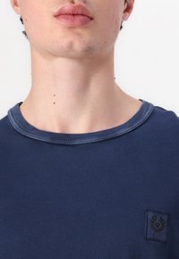 Námorně modré tričko s kulatým výstřihem, žebrovaným límcem a malou nášivkou na levé straně. Tkanina vypadá hladce s mírným leskem.