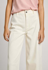 Pantaloni a gamba larga in crema chiara realizzati in cotone, con chiusura a bottoni, vestibilità rilassata e due tasche laterali. Tessuto liscio e texturizzato.