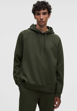 Joven con cabello rizado que lleva una sudadera con capucha verde oscuro y pantalones deportivos a juego, de pie con una mano en el bolsillo.