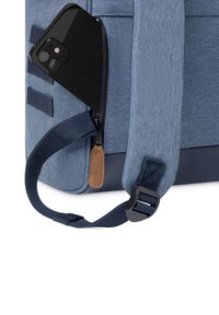 Zaino in tessuto blu con una cerniera scura, con uno smartphone nero parzialmente inserito. Dettaglio in pelle marrone sul tirazip.