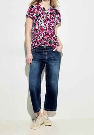 Kobieta ubrana w jaskrawą różową, czarną i purpurową koszulkę z krótkim rękawem w wzory, ciemnoniebieskie krótkie dżinsy i beżowe trampki, stojąca przy białej ścianie.