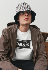 Brun lynlåsjakke over en hvid t-shirt med en sort rektangulær grafik, parret med en ternet bøttehat i blå og brun.