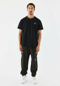 Camiseta negra con logo en el pecho y un cuello estampado, combinada con pantalones negros de corte holgado que tienen acentos blancos y rayas laterales, usados con zapatillas blancas.