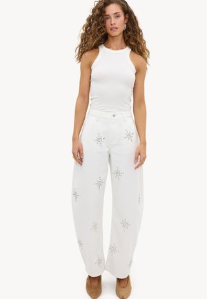 Vrouw met krullend haar die een witte mouwloze top en wijde witte broek met stervormige versieringen draagt, lichtbruine puntige schoenen.