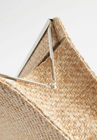 Primer plano de un bolso de fibra natural tejido con marco y bisagra de metal plateado sobre un fondo blanco.
