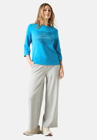 Hellblauer Sweatshirt mit Dreiviertelärmeln und geometrischem Druck, kombiniert mit hellgrauen weiten Hosen und beigen Sneakers.