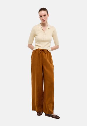 Femme debout, mains derrière le dos, portant un haut en maille beige à manches courtes, un pantalon pattes d'éléphant rouille et des ballerines tressées marron foncé.