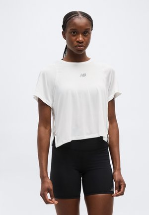 Kvinde med fletninger iført en løs hvid New Balance T-shirt og tætsiddende sorte atletikshorts mod en ensfarvet baggrund.