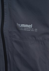 Close-up van donkere stof met "hummel Techwear - Ontworpen door HML Performance Sportswear Sinds 1923" gedrukt in lichtgrijze tekst.
