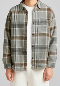 Camisa de franela a cuadros grises con detalles en naranja y blanco, con botón delantero y un bolsillo en el pecho. Se combina con un suéter blanco debajo.