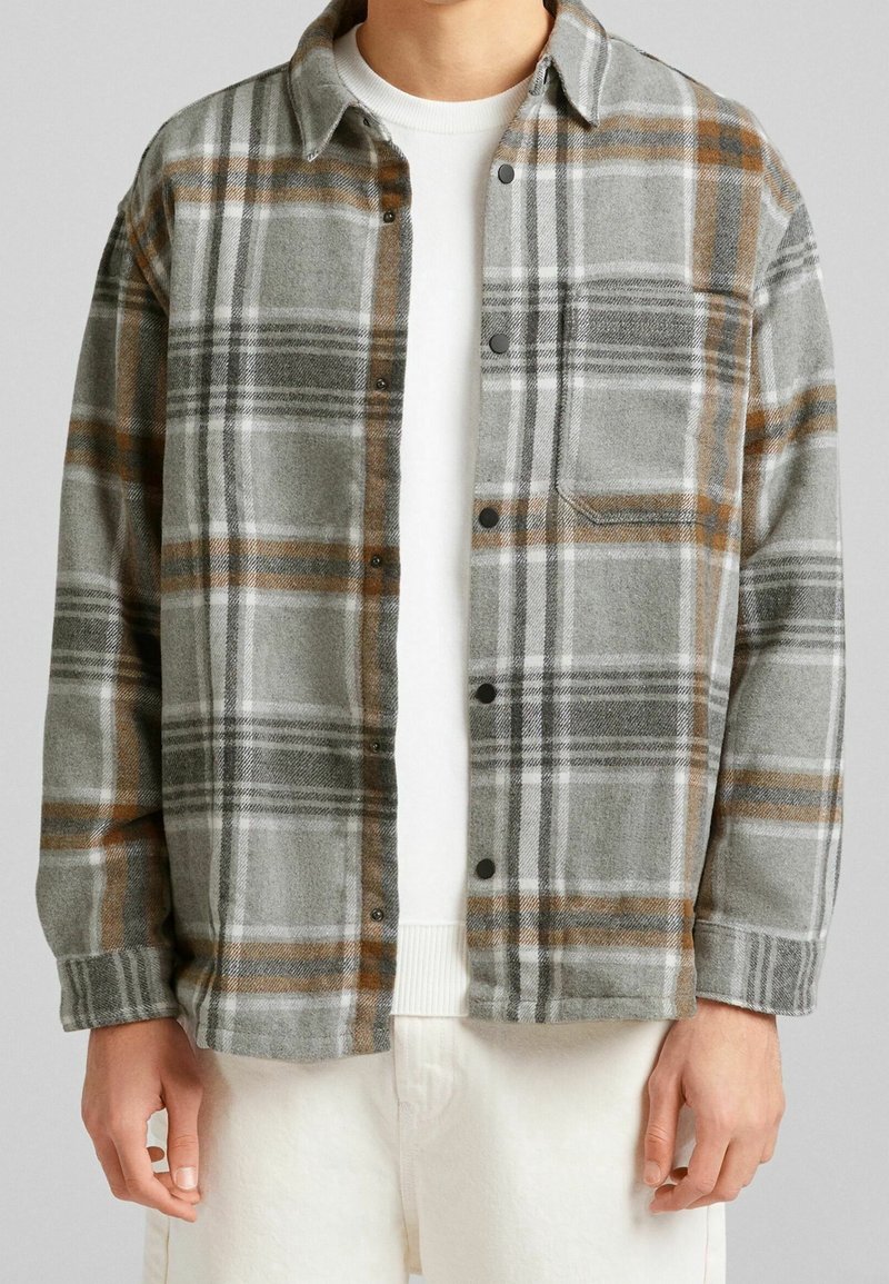 Camisa de franela a cuadros grises con detalles en naranja y blanco, con botón delantero y un bolsillo en el pecho. Se combina con un suéter blanco debajo.
