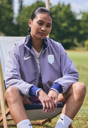 Lys lilla zip-up jakke med blå detaljer og en logo, brukt over en hvit skjorte og mønstrede shorts. Avslappet passform med ribbestrikkede mansjetter.
