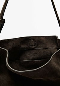 Mørkebrune ruskinds tote-tasker med en blød tekstur, udstyret med en cirkulær knaplukning indeni og et minimalistisk design. Indersiden viser et uforet rum.