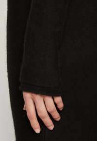 Black knitted sweater sleeve, featuring a wide cuff and textured fabric. A hand rests on the sleeve, showing the garment's softness.