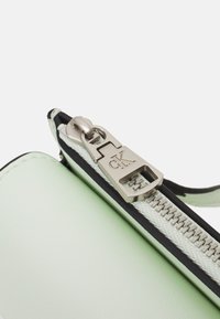 Róg zielonej skórzanej torby z srebrnym zamkiem błyskawicznym, czarną lamówką i tkaninową zawieszką, z wytłoczonym logo na zamku.