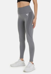 Grijze high-waisted leggings met een zachte textuur en een accent van zijpanelen, met een wit logo op de bovenbeen. Gecombineerd met witte sportschoenen.