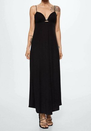 Kobieta w czarnej, długiej sukience na ramiączkach, z cienkimi paskami, czarne sandały z zapięciem na sznurowadła oraz widoczne kwiatowe tatuaże na ramionach i barku.