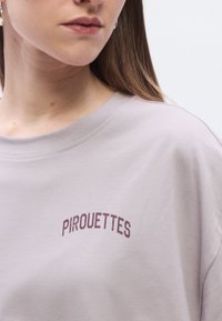 Light lavender t-shirt with a relaxed fit, featuring maroon "PIRROUETTES" text above the chest area and a smooth cotton texture.