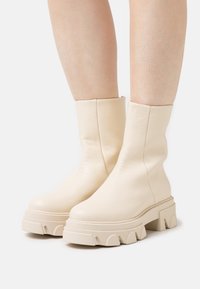 Cremfarbene Leder-Stiefel bis zum Knöchel mit glatter Textur, ausgestattet mit einer dicken, klobigen Sohle mit wellenförmigem Design und minimalen Nähdetails.