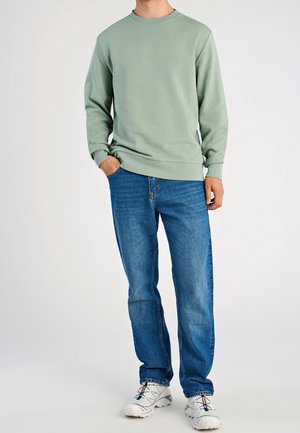 Homme portant un sweat-shirt vert clair, un jean bleu et des baskets blanches, debout avec une main dans la poche devant un fond uni.