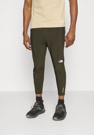 Mann trägt beige Hemd, dunkelolivfarbene, zulaufende Hose mit The North Face Logo, schwarze Socken und schwarze Wanderschuhe mit grauen Schnürsenkeln.