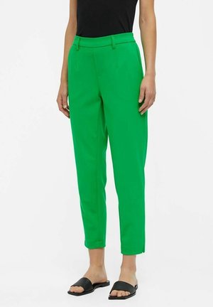 Frau trägt hoch taillierte, leuchtend grüne, konisch zulaufende Hose mit Seitentaschen und schwarzen Slide-Sandalen, steht vor weißem Hintergrund.