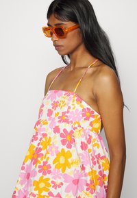Vestido de verão floral com flores rosas e amarelas sobre um fundo branco, apresentando alças finas e um design solto e fluido. Óculos de sol laranja.