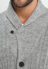 Grå strikket cardigan med sjalskrage, to svarte knapper og et teksturert kabelstrikkmønster. Mykt stoff med en koselig utseende.