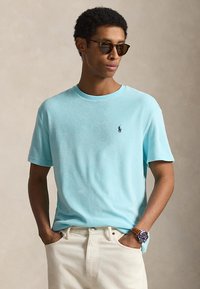 Άνδρας ντυμένος με κοντομάνικο γαλάζιο πουκάμισο, μπεζ παντελόνι, καφέ ρολόι χειρός και σκούρα γυαλιά ηλίου, στέκεται με τα χέρια στις τσέπες.