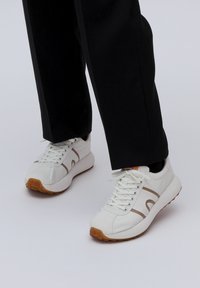 Λευκά αθλητικά παπούτσια με καφέ λεπτομέρειες, σχέδιο με κορδόνια, ελαστική σόλα και λεία υφή δέρματος, τοποθετημένα με μαύρα παντελόνια.