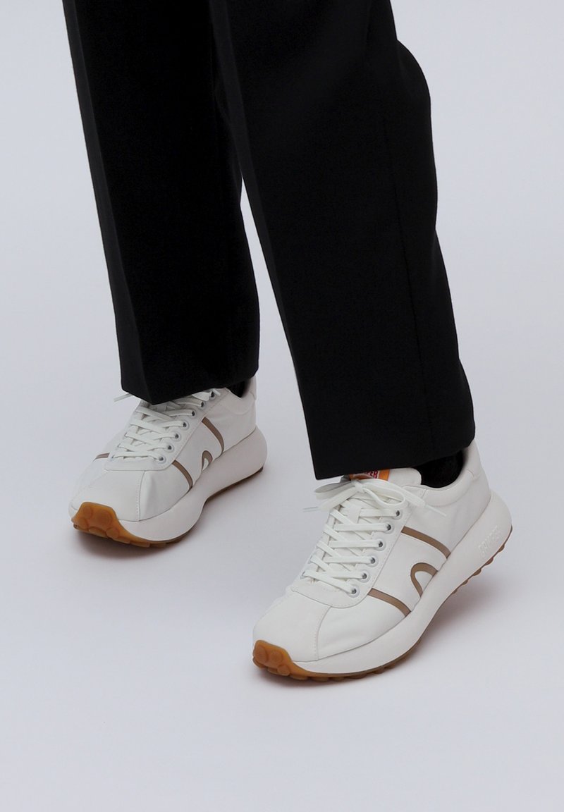 Λευκά αθλητικά παπούτσια με καφέ λεπτομέρειες, σχέδιο με κορδόνια, ελαστική σόλα και λεία υφή δέρματος, τοποθετημένα με μαύρα παντελόνια.