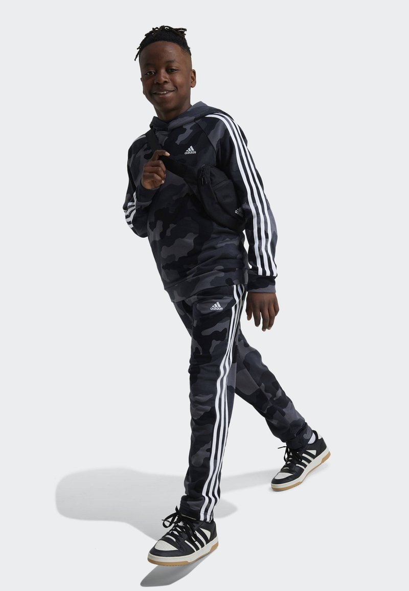 Tuta mimetica nera e grigia con strisce bianche. Il modello indossa una borsa a tracolla nera e sneakers nere con dettagli bianchi.