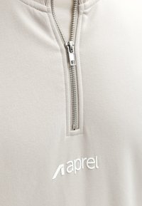 Primer plano de una sudadera beige claro de media cremallera con cremallera plateada y el logo "aprel" en texto blanco debajo de la cremallera.