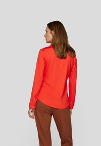 Long-sleeve shirt in bright orange fabric, featuring a pointed collar, button cuffs, and a curved hem, paired with brown trousers.